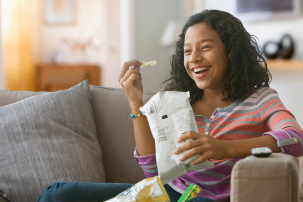 hispanic girl eating potato chips on sofa - junk food stock pictures, royalty-free photos & images