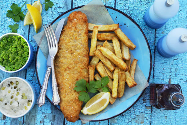 image of fish and chips (cod) garnished with parsley on turquoise plate with greaseproof parchment, knife and fork, lemon slice, bowls containing tartare sauce and mushy peas, bottle of vinegar, turquoise background, elevated