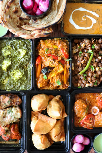 image of indian takeaway dishes in black plastic, single-use food containers, kadai chicken, chicken bhuna, palak paneer, samosas, murgh malai tikka, pink pickled onions, mint and coriander dip, lachha paratha, pindi chole a