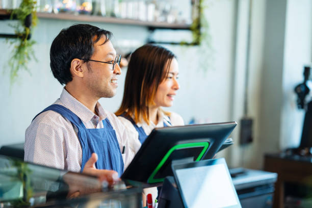 improve and increase the customer experience in coffee shops. shot of a male japanese coffee shop owner and a female waitress at a cashier counter and greeting customers as they enter the coffee shop. - junk food stock pictur