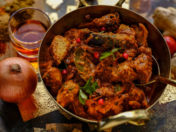 indian chicken vindaloo in a brass wok ready to be served, on a grunge metal, wood and gold leaf background with fresh vegetables, spices and red wine vinegar, ingredients of the dish, accompanying the scene. - food stock pic