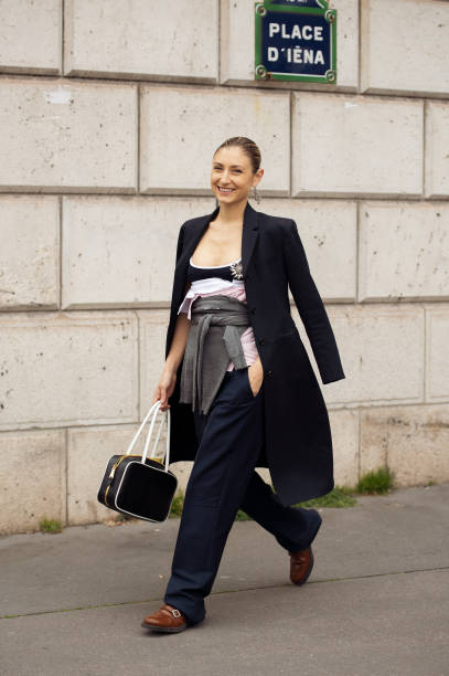 Jenny Walton wears a black and white Miu Miu leather bag, a black long coat, a grey Miu Miu sweater around the waist, a black and white Miu Miu tank...
