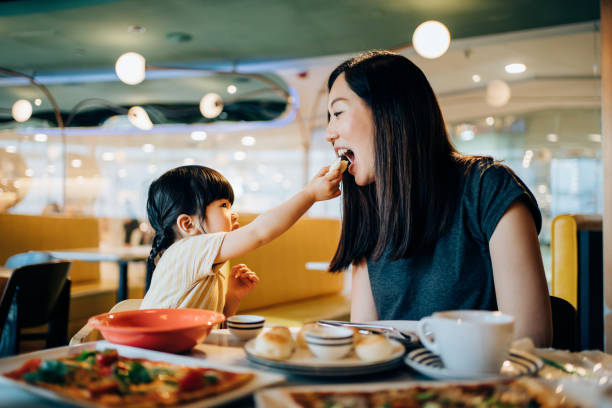 joyful young asian mother and lovely little daughter enjoying lunch together in restaurant. they are sharing food, enjoying family bonding time and a happy meal at the dining table. family and eating out lifestyle - food stoc