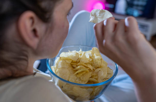 June 2023, Saxony, Leipzig: ILLUSTRATION - A woman is eating chips in a living room in front of the TV. Photo: Hendrik Schmidt/dpa