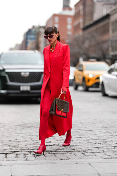 Katya Tolstova wears dark red sunglasses, gold hoop earrings, gold necklaces, bright cherry red blazer jacket, bright cherry red long sleeve sweater,...