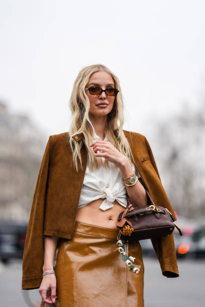 Leonie Hanne wears dark brown sunglasses, white collar tied Miu Miu shirt, dark brown unzipped Miu Miu suede jacket, gold Cartier Bracciale Juste...