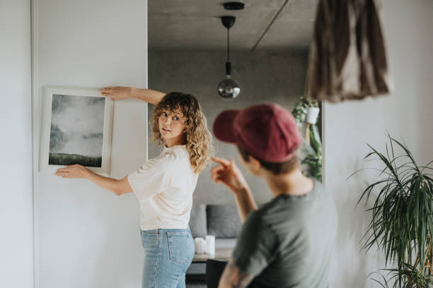 lesbian woman taking advice from girlfriend while hanging frame on wall at home - home decoration stock pictures, royalty-free photos & images