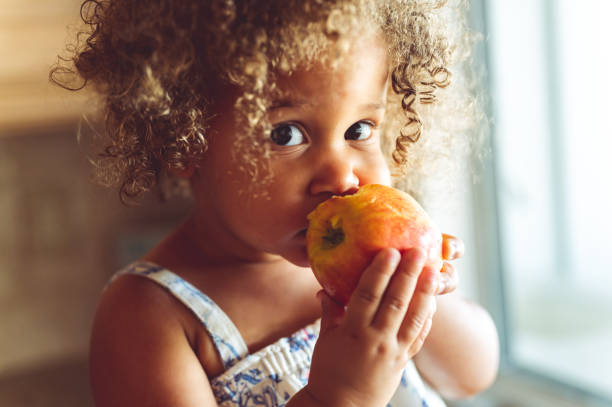 linda niña de raza mixta comiendo una manzana roja fresca, comiendo saludable en casa - food fotografías e imágenes de stock