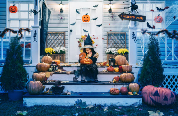 little girl in witch costume sitting on the stairs in front of the house and holding jack-o-lantern pumpkins on halloween trick or treat - home decoration stock pictures, royalty-free photos & images