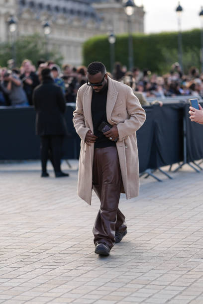 Maitre Gims wears black sunglasses, light brown blazer jacket, shiny dark brown leather pants, dark brown leather shoes, silver rings, outside Louis...