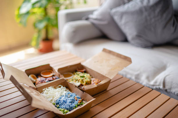 malaysian food nasi kerabu, nasi ulam and ayam percik in recycled paper container with sauce on coffee table beside sofa in living room - junk food stock pictures, royalty-free photos & images
