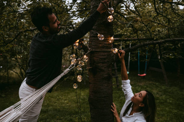male and female friends hanging lighting equipment in backyard during dinner party - garden decoration stock pictures, royalty-free photos & images