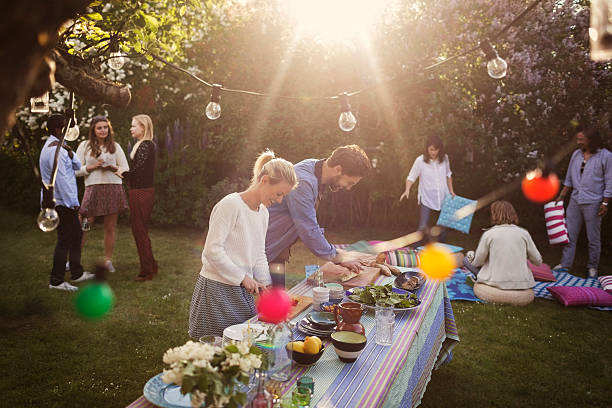 man and woman preparing food at dining table with friends in background during summer party - garden decoration stock pictures, royalty-free photos & images