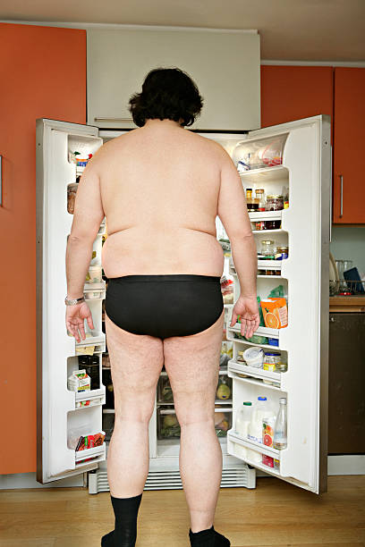 man wearing underwear, standing by open fridge, rear view - junk food stock-fotos und bilder