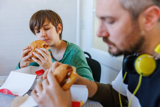 man with headphones and his preadolescent son having take out burgers at the table - junk food stock pictures, royalty-free photos & images