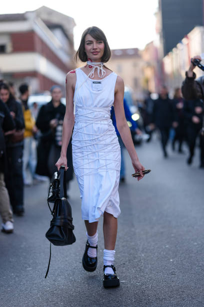 Mara LaFontan wears cream cloth wrapped choker necklace, white tied wrapped sleeveless Moschino midi dress, shiny black Moschino leather bags, white...