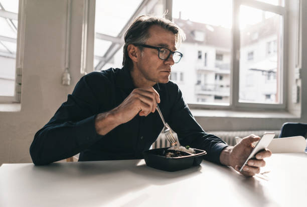 mature businessman checking cell phone during lunch break in office - junk food stock pictures, royalty-free photos & images
