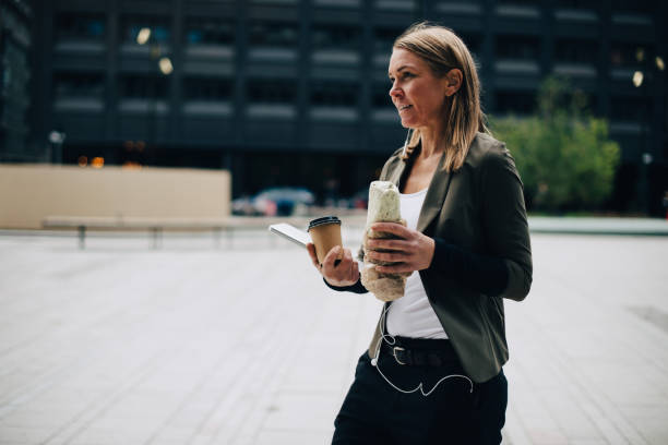 mature businesswoman carrying food and drink with laptop while walking on footpath in city - junk food stock pictures, royalty-free photos & images