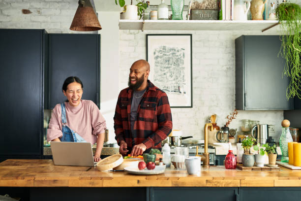 mature chinese woman and black mid adult man preparing food and laughing in kitchen preparing food with laptop on worktop - food stock pictures, royalty-free photos & images