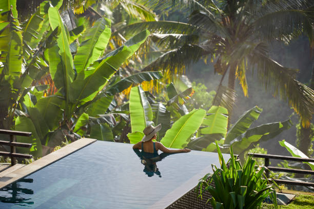 mature fit asian woman relaxing on edge of infinity pool, admiring jungle view with early morning mist - travel stock pictures, royalty-free photos & images