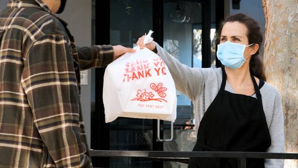 mature restaurant female worker delivering a bag with meal at a restaurant curbside pickup wearing a protective face mask - junk food stock pictures, royalty-free photos & images