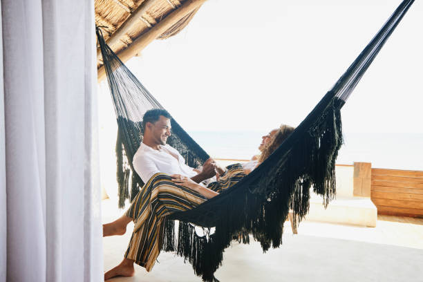 medium wide shot of smiling couple holding hands while sitting in hammock on deck of luxury tropical villa - travel stock pictures, royalty-free photos & images