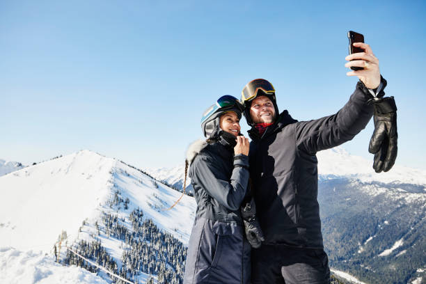 medium wide shot of smiling couple taking selfie while standing at the top of ski resort on winter morning - travel stock pictures, royalty-free photos & images