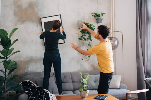 mid adult man guiding girlfriend in hanging picture frame on wall at new home - home decoration stock pictures, royalty-free photos & images