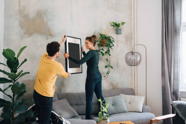 mid adult man passing picture frame to girlfriend against wall at new home - home decoration stock pictures, royalty-free photos & images