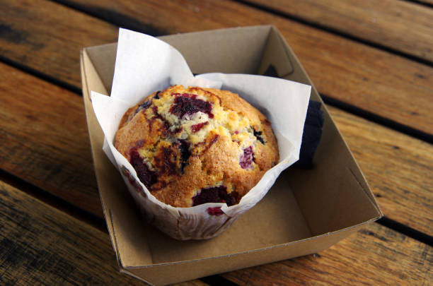 mixed berry, white chocolate and coconut muffin in waxed paper in a cardboard tray on a wooden outdoor table - junk food stock pictures, royalty-free photos & images