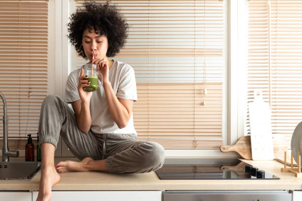 mixed race black latina woman drinking healthy green juice sitting on kitchen counter. copy space. healthy eating. - food stock pictures, royalty-free photos & images