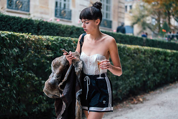 Model Kye Howell checks her phone, drinks champagne, and wears a white corset top and black sporty Adidas shorts after the Dior show at Musee Rodin...