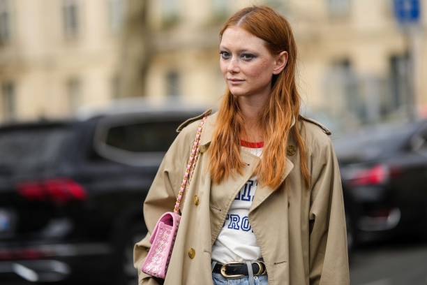 Model wears a beige long belted trench coat, blue denim pants, a pink and white checkered print pattern tweed shoulder bag from Chanel, a black shiny...