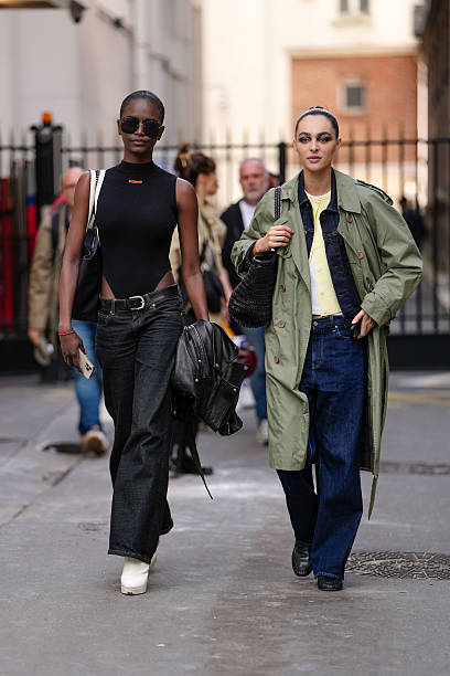 Model wears a sleeveless black top with a high neckline, paired with high-waisted dark denim jeans. Accessories include large black sunglasses and a...