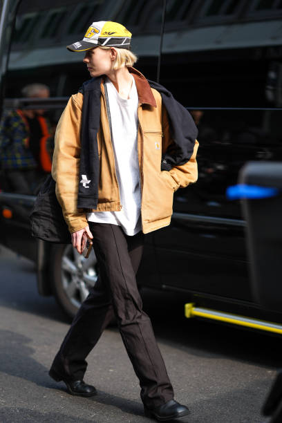 Model wears a white t-shirt, a beige leather jacket, a scarf, a black white and yellow cap, black pants, black leather shoes, outside Onitsuka Tiger...