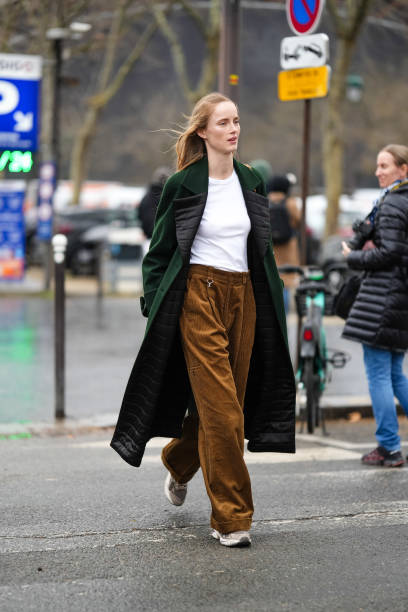 Model wears a white t-shirt, a green long coat with black quilted lapels, brown flared corduroy pants, sneakers shoes, outside Chanel, during the...