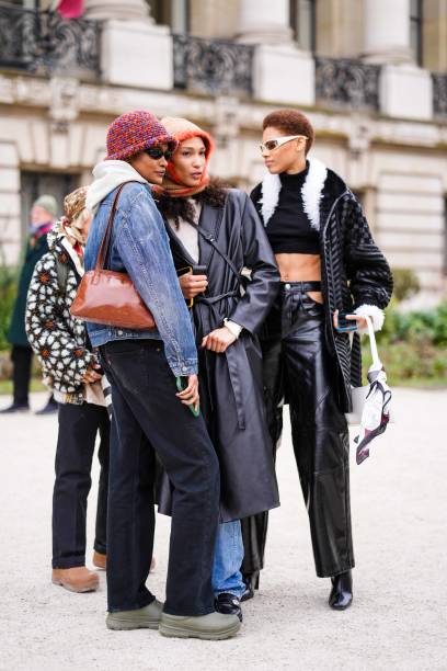 Model wears black sunglasses, a white hoodie sweater, a blue faded denim jacket, a camel shiny varnished leather shoulder bag, black denim pants,...