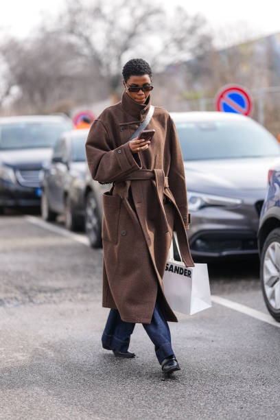 Model wears black sunglasses, silver earrings, dark brown wool oversized coat, a gold ring, black crossbody leather bag, dark navy blue lose denim...