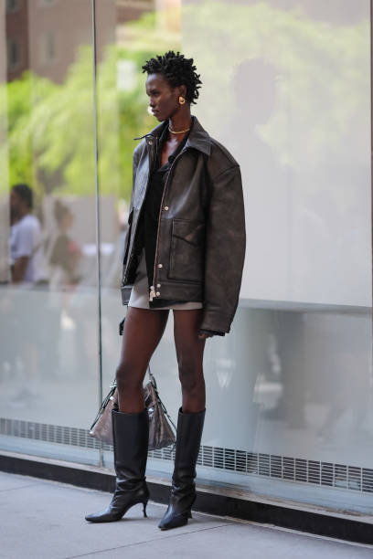 Model wears gold earrings, gold necklace, black shirt, dark brown oversized blazer jacket in leather, light gray mini skirt, shiny black pointed toe...