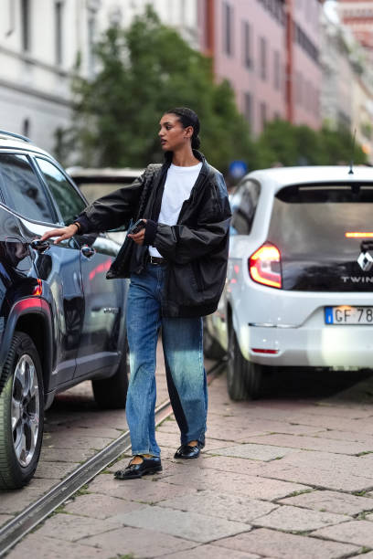 Model wears white cotton t-shirt, black oversized baggy leather jacket, shiny black woven leather bag, black leather belt, light blue denim jean...