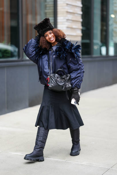 Naomi Elizee wears black knit hat, dark blue oversized puffer bomber jacket, shiny black leather bag, shiny black leather gloves, black midi skirt,...