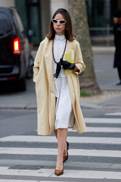 Natasha Goldenberg wears white blouse, white midi skirt, yellow coat, brown heels, black leather gloves, white sunglasses, outside Miu Miu, during...