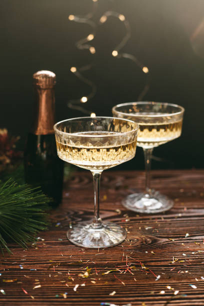 new year greeting card. champagne vintage glasses and bottle decorated with christmas decorations, pine, lights and confetti over dark wood background - home decoration stock pictures, royalty-free photos & images
