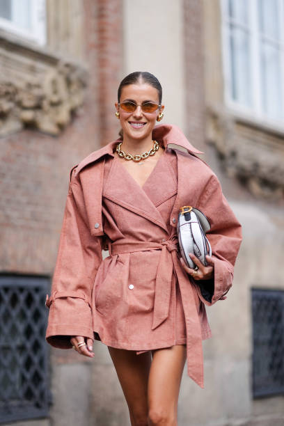 Nina Sandbech wears light brown sunglasses, gold earrings, gold chain necklace, sand beige oversized blazer belted mini trench dress, white and black...