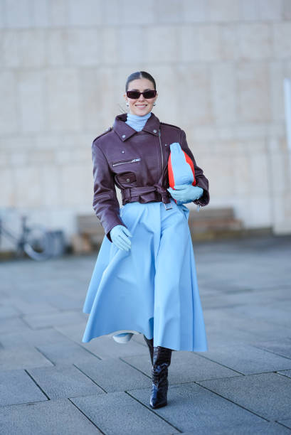 Nina Suess wears sunglasses , a blue turtleneck long gathered oversized dress, a purple leather biker jacket , a red and blue bag, black leather...