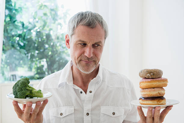 older caucasian man choosing between broccoli and donuts - junk food stock pictures, royalty-free photos & images