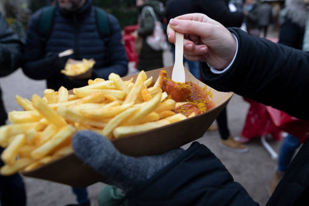 over the shoulder view of woman eating chips and curry sausage from a paper tray at a german christmas market. - junk food stock pictures, royalty-free photos & images