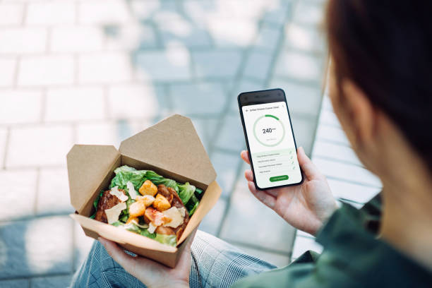 over the shoulder view of young asian businesswoman using fitness plan mobile app on smartphone to tailor make her daily diet meal plan, checking the nutrition facts and calories intake of the food while having meal outdoors 