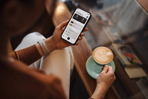 over the shoulder view of young asian woman managing personal banking and finance with online banking app on smartphone, sitting on sofa at home. financial planning. investment. wealth management, banking and finance concept.