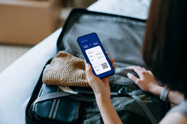 over the shoulder view of young asian woman using smartphone, checking digital flight ticket and boarding pass on device screen while packing a suitcase on bed for a trip. planning for travel. travel and vacation concept - tr